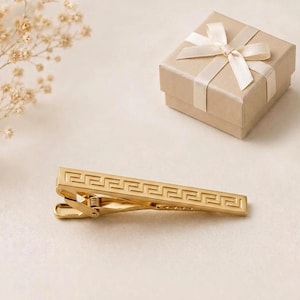 May include: A gold-toned metal tie clip featuring a repeating geometric Greek key pattern engraved along the front surface. The tie clip is positioned horizontally next to a square, light brown gift box tied with a cream-coloured satin ribbon.