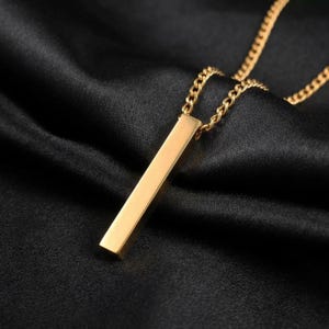 Op de afbeelding: Een goudkleurige metalen ketting met een lange, rechthoekige staafhanger die aan een bijpassende goudkleurige ketting hangt. De hanger heeft een glad, gepolijst oppervlak met strakke, rechte randen en hangt verticaal aan de ketting.