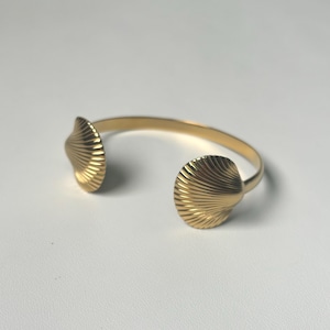 Peut inclure: Un bracelet manchette doré doté de deux extrémités symétriques en forme de coquillage. La surface métallique du bracelet est lisse et polie, tandis que les deux embouts en forme de coquille présentent des stries rayonnantes détaillées.