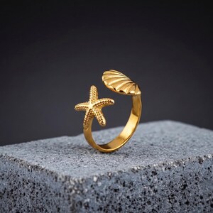 Puede incluir: Un anillo abierto de tono dorado que presenta una estrella de mar texturizada en un extremo y un diseño de concha marina estriada en el otro. La banda es lisa y circular, elaborada con un acabado metálico pulido que refleja la luz.