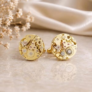 May include: A pair of round gold-toned cufflinks featuring exposed mechanical watch movements. Each cufflink displays intricate brass-colored gears, small silver-toned screws, and tiny pink synthetic rubies set within the circular metal frame.