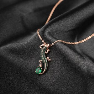 Op de afbeelding: Een roségouden ketting met een hagedisvormige hanger die op de rug is ingelegd met kleine groene steentjes. De staart van de hagedis krult naar beneden en houdt een enkele, ovaal geslepen groene edelsteen vast aan een fijne ketting.
