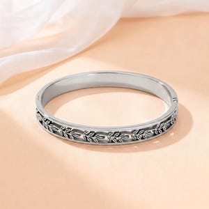 May include: A silver-toned metal bangle bracelet featuring a continuous engraved leaf pattern around the outer band. The bracelet has a smooth interior surface and a hinged clasp mechanism visible on the side, showcasing a polished finish.