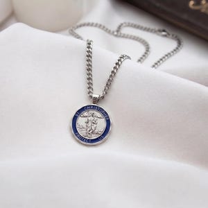 May include: A silver-toned metal pendant necklace featuring a circular St. Christopher medallion with a deep blue enamel border. The centre depicts a raised relief figure carrying a child, with the text ST. CHRISTOPHER PROTECT US inscribed.