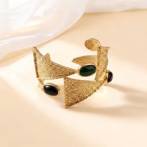 Può includere: Un bracciale a fascia color oro caratterizzato da una superficie geometrica testurizzata con pannelli triangolari. Il gioiello è impreziosito da tre pietre cabochon ovali e lisce, tra cui una verde scuro e due nere, incastonate nel metallo.