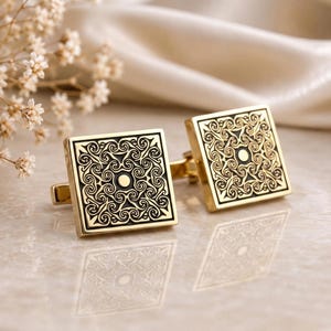 May include: A pair of square gold-toned cufflinks featuring a black recessed background with an intricate, symmetrical swirling filigree pattern and a small circular center detail. The metal surfaces have a polished finish that reflects light.