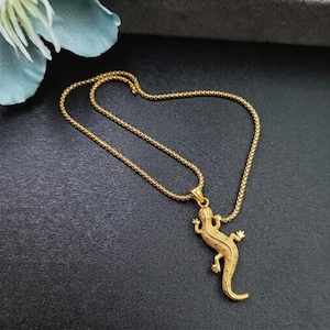Pode incluir: Um colar de tom dourado com um pingente em forma de lagarto texturizado suspenso por uma corrente fina de elos quadrados. O pingente exibe escamas detalhadas e uma cauda curva, enquanto a corrente é fina e uniforme em sua estrutura.