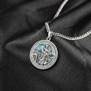 May include: A silver-toned circular pendant necklace featuring an embossed relief of Saint Christopher carrying a child. The central figure is set against an iridescent abalone shell inlay. The border is inscribed with the text SAINT CHRISTOPHER PROTECT US.
