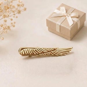 May include: A gold-toned metal hair clip shaped like a single wing with detailed feather engravings, positioned next to a square cardboard gift box tied with a cream-coloured satin ribbon bow and a small sprig of dried baby's breath flowers.