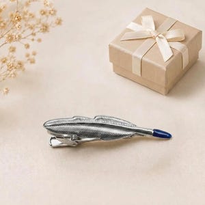 May include: A silver-toned metal hair clip shaped like a feather with detailed etched lines and a small, smooth, dark blue accent at the pointed tip. The clip features a standard spring-loaded mechanism on the underside for securing hair.