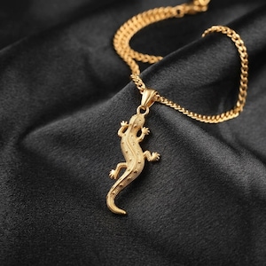 May include: A gold-toned necklace featuring a textured lizard pendant suspended from a matching curb chain. The lizard charm displays detailed scales and a curved tail, with the entire piece crafted in a polished metallic finish that catches light.