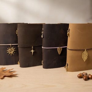 Pode incluir: Quatro diários de couro em vários tons de castanho, cada um preso com uma banda elástica colorida. Os diários apresentam berloques de metal dourado, incluindo um sol, uma estrela, um coração e uma folha, presos aos fechos elásticos.