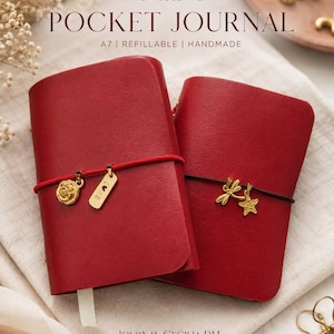 Puede incluir: Dos libretas de bolsillo A7 de cuero rojo hechas a mano. La libreta izquierda tiene una banda elástica roja con dijes dorados de rosa y un rectángulo con la palabra "LOVE". La derecha tiene una banda elástica negra con dijes de libélula y estrella.