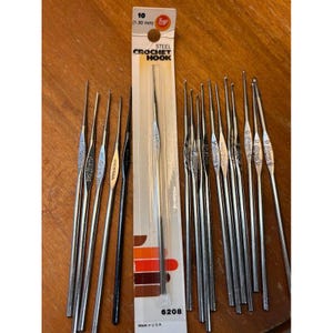 May include: A collection of thin steel crochet hooks, including one packaged in a white card labeled 10 (1.30 mm) STEEL CROCHET HOOK. The hooks are metallic silver, with one black hook, and feature various stamped size markings along their shafts.