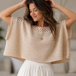 May include: A beige crochet top featuring a wide, boxy silhouette with short sleeves and an intricate openwork pattern around the neckline. The garment is crafted from a light-colored yarn and is paired with a white skirt featuring a drawstring waist.