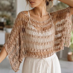 Può includere: Un top all'uncinetto color beige caratterizzato da un motivo a maglia aperta con fasce orizzontali di punti pieni. Il capo presenta una silhouette ampia e morbida con maniche corte e scollo rotondo, indossato sopra una canotta bianca.