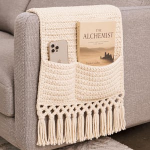 May include: A cream-colored crocheted armrest organizer featuring two pockets, a decorative knotted fringe at the bottom, and a textured stitch pattern. The pockets contain a smartphone and a copy of the book The Alchemist by Paulo Coelho.