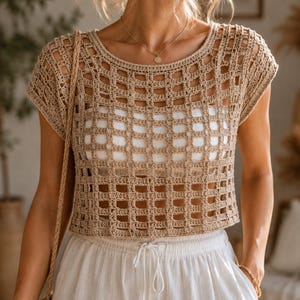 Puede incluir: Un top de ganchillo beige con un patrón de rejilla abierto, mangas cortas y cuello redondo. La prenda se lleva sobre una capa base blanca sólida y se combina con pantalones blancos con cordón, creando un aspecto texturizado y superpuesto.