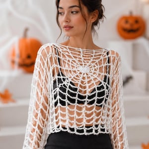 May include: A white long-sleeve crochet top featuring an open-knit spiderweb pattern. The garment has a wide neckline and a cropped silhouette, worn over a black bra and a black skirt. The intricate web design radiates from the centre chest.