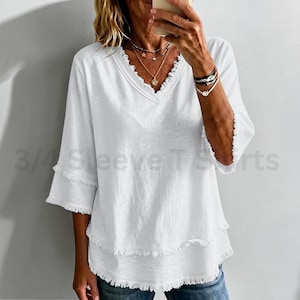 May include: A white V-neck top featuring three-quarter length sleeves with frayed edges and a layered hemline with matching frayed detailing. The garment includes the text 3/4 Sleeve T-Shirts overlaid on the image. The fabric has a textured appearance.