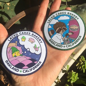 May include: Two round embroidered patches with the text "Laced Cakes Bakery Oakland California" and a cartoon image. One patch features a raccoon painting a pie and the other features a sleeping character dreaming of cakes.