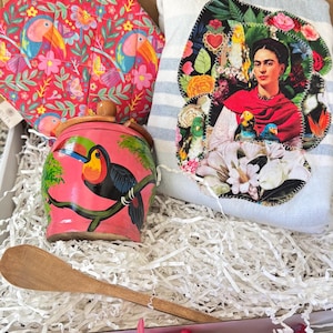 May include: A gift box containing a pink quilted potholder with a toucan and floral print, a small pink wooden jar with a toucan illustration, a light blue and white striped cloth with a Frida Kahlo portrait patch, and a wooden spoon.