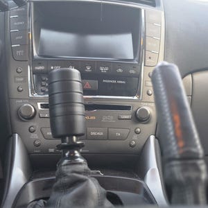 May include: A black cylindrical gear shift knob with horizontal grooves, mounted on a black leather boot within a vehicle centre console. The console features a digital display, climate control buttons, and a Mark Levinson audio system interface.