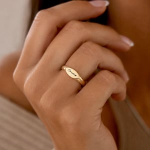 Può includere: Un anello color oro lucido caratterizzato da una superficie di forma ovale con il nome Christine inciso in un carattere corsivo nero. La fascia è liscia e arrotondata, mantiene una larghezza costante attorno al dito e mostra un design minimalista.