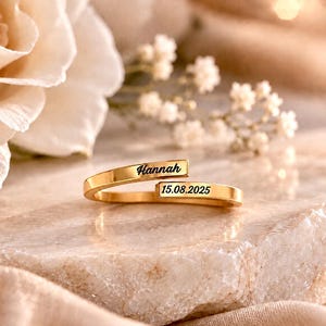 Peut inclure: Une bague ouverte dorée composée de deux bandes plates et parallèles. Une bande est gravée du prénom Hannah dans une police cursive noire, et l'autre bande porte la date 15.08.2025 inscrite dans une police noire sans empattement.