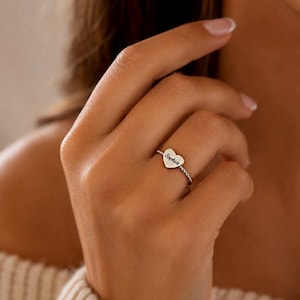 Op de afbeelding: Een zilverkleurige ring met een kleine hartvormige bedel waarop de naam Sophia is gegraveerd. De ringband heeft een gedraaide, touwachtige textuur, wat zorgt voor een gedetailleerd uiterlijk. Het metaal heeft een gepolijste afwerking.