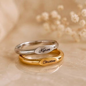 Könnte beinhalten: Zwei übereinanderliegende Siegelringe mit glatten, polierten Ringschienen. Der obere Ring ist silberfarben und trägt den Namen Karen in schwarzer Schreibschrift. Der untere Ring ist goldfarben und trägt den Namen Christine in schwarzer Schrift.