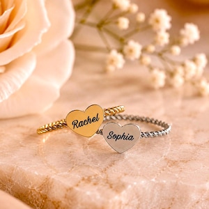 Pode incluir: Dois anéis com bandas torcidas, um em tom dourado e outro em tom prateado. Cada anel apresenta um pingente plano em forma de coração gravado com um nome em letra cursiva preta. O anel dourado tem o nome Rachel e o prateado Sophia.