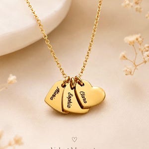 Könnte beinhalten: Eine goldfarbene Halskette mit drei herzförmigen Anhängern an einer feinen Kette. Jedes Herz ist mit einem Namen graviert: Maria, Sophia und Clara. Der Text Velvet Moments Personalized Jewelry steht unten im Bild.