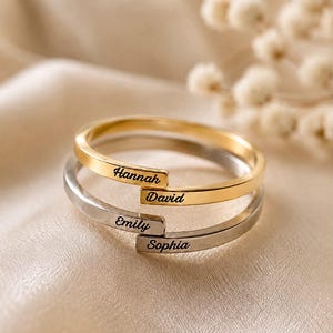 Può includere: Un set di anelli impilabili composto da due fasce, una color oro e una color argento. La fascia dorata è incisa con i nomi Hannah e David, mentre la fascia argentata riporta i nomi Emily e Sophia in un carattere corsivo di colore nero.