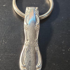 May include: A silver-toned metal keychain featuring a handle from a piece of flatware. The handle has an embossed floral pattern with curved lines and a smooth, polished center section. A circular metal split ring is attached to the top end.