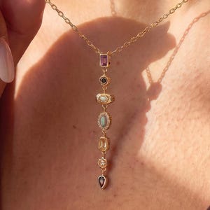 May include: A gold chain necklace featuring a vertical drop pendant composed of seven small, multi-colored gemstones in various shapes, including rectangular, round, oval, and teardrop settings, suspended from a delicate gold link chain.
