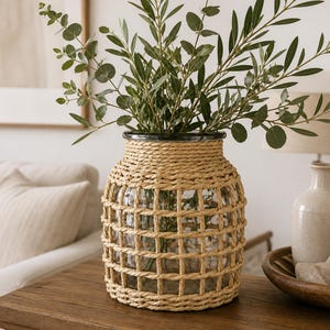 Op de afbeelding: Een vaas van helder glas omwikkeld met een dik, beige touwnet dat een rasterpatroon vormt aan de buitenkant. De vaas heeft een brede, ronde vorm met een smallere hals en is gevuld met groene takken met kleine, ovale bladeren.