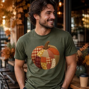 May include: An olive green short-sleeve t-shirt featuring a large, central graphic of a pumpkin constructed from various patchwork fabric patterns, including red and white gingham, orange polka dots, and small floral prints in warm autumn tones.