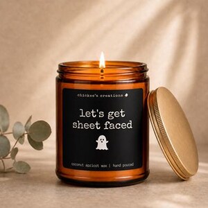 Peut inclure: Une bougie allumée dans un pot en verre ambré avec une étiquette noire portant le texte 'chickee's creations', 'let's get sheet faced', une icône de fantôme blanc et 'coconut apricot wax | hand poured'. Un couvercle en métal doré est posé à côté.
