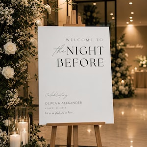 Può includere: Un cartello di benvenuto per matrimonio rettangolare bianco su un cavalletto di legno con testo nero che recita: WELCOME TO the NIGHT BEFORE, Celebrating OLIVIA & ALEXANDER, AUGUST 21, 2027, We're so glad you're here. Design pulito ed essenziale.