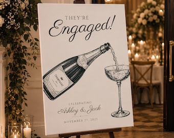 May include: A white rectangular sign on an easel featuring the black text "THEY'RE Engaged!" above a detailed black and white illustration of a champagne bottle pouring into a coupe glass, with "CELEBRATING ASHLEY & JACK" and "NOVEMBER 27, 2025".