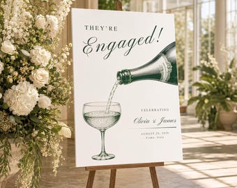 May include: A white rectangular sign on a wooden easel featuring the text "THEY'RE Engaged!" above an illustration of a dark green champagne bottle pouring into a glass. Below, the text reads "CELEBRATING Olivia & James AUGUST 20, 2026 Como, Italy."