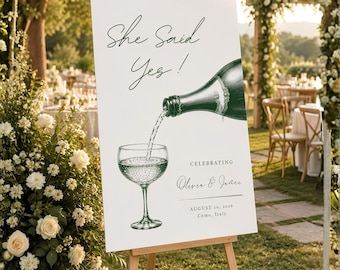 May include: A white rectangular wedding sign on a wooden easel featuring a dark green illustration of a champagne bottle pouring into a glass. The text reads She Said Yes!, CELEBRATING, Olivia & James, and AUGUST 20, 2026, Como, Italy.