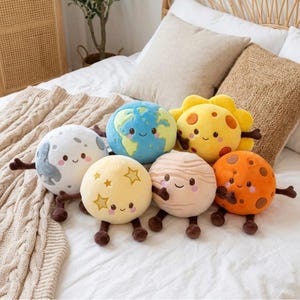 May include: A collection of six plush toys shaped like celestial bodies, including a blue and green Earth, a yellow sun, a white moon, a pale yellow star-patterned sphere, a beige striped planet, and an orange cratered planet, all with brown limbs.