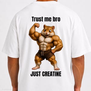 Può includere: Una t-shirt bianca con la grafica di un criceto marrone muscoloso che flette le braccia. Sopra il criceto, il testo recita "Trust me bro" e sotto il criceto, il testo recita "JUST CREATINE" in lettere maiuscole nere e in grassetto.