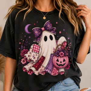 May include: A black short-sleeved t-shirt featuring a graphic of a white ghost wearing a large purple floral bow. The ghost is surrounded by pink and purple flowers, a crescent moon, stars, and is holding a pink jack-o'-lantern bucket.