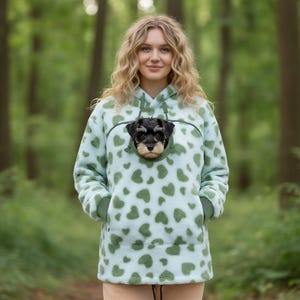 May include: A light blue fleece hoodie featuring a repeating pattern of dark green hearts. The garment includes a specialized front pouch designed to carry a small dog, with the head of a black and tan puppy visible through the circular opening.