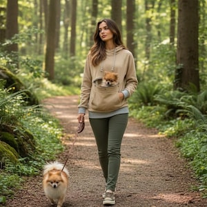 May include: A tan hooded sweatshirt featuring a large front pouch pocket containing a small brown dog, paired with olive green pants. A second small brown dog is walking on a leash on a dirt path in a forest setting with green trees and ferns.