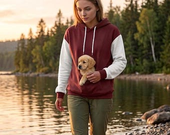 Puede incluir: Una sudadera con capucha color granate con mangas blancas y un bolsillo frontal que contiene un pequeño cachorro dorado. La sudadera tiene capucha con cordón y puños acanalados. Se combina con pantalones verde oliva y botas de montaña.