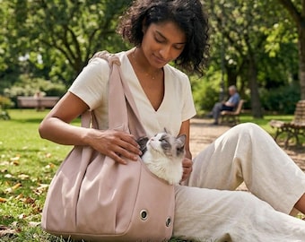 Puede incluir: Un bolso transportín para mascotas de color rosa empolvado con una correa ancha para el hombro y dos ojales metálicos circulares de ventilación en la parte inferior frontal. Un gato de pelo largo blanco y gris descansa dentro del bolso.